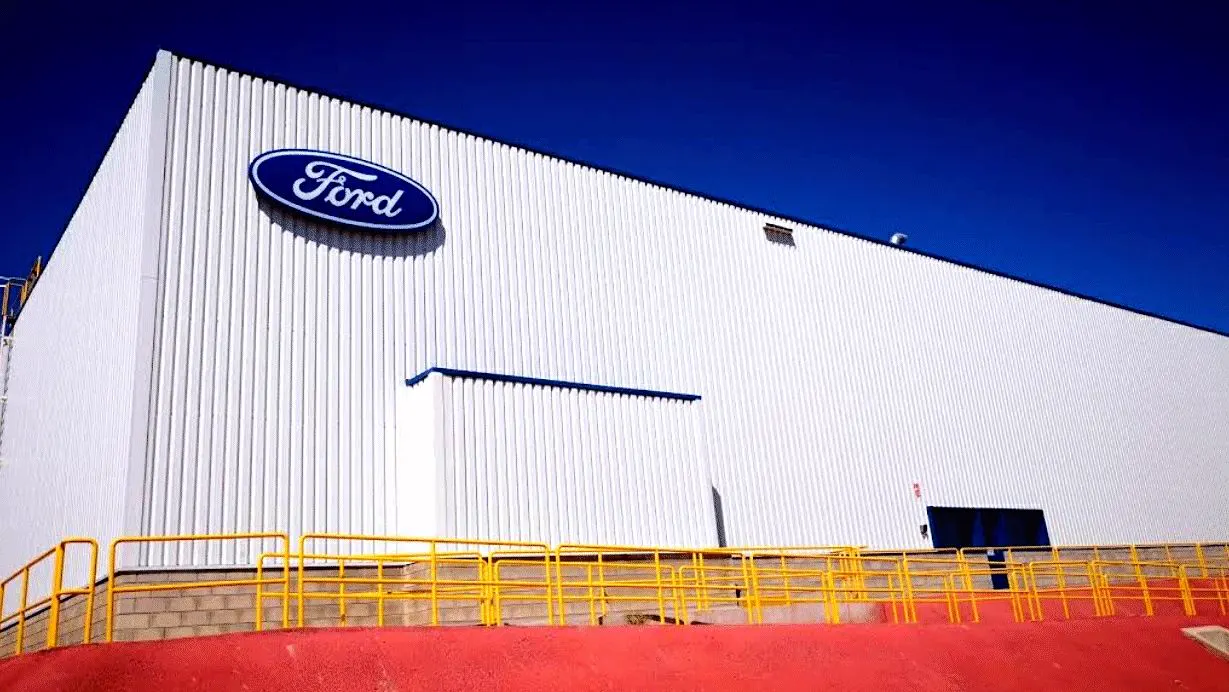 Fachada de una nave blanca de la fábrica de Ford en Almussafes con el logotipo azul de Ford y una barandilla amarilla en primer plano.