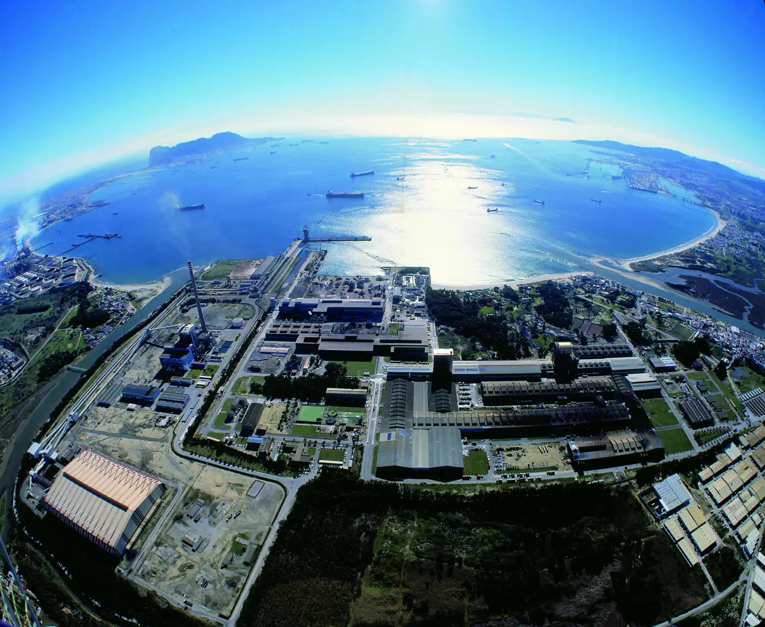 Vista aérea de un gran complejo siderúrgico con naves industriales y una chimenea junto al mar, en la bahía de Algeciras, con decenas de buques fondeados y un promontorio rocoso al fondo.