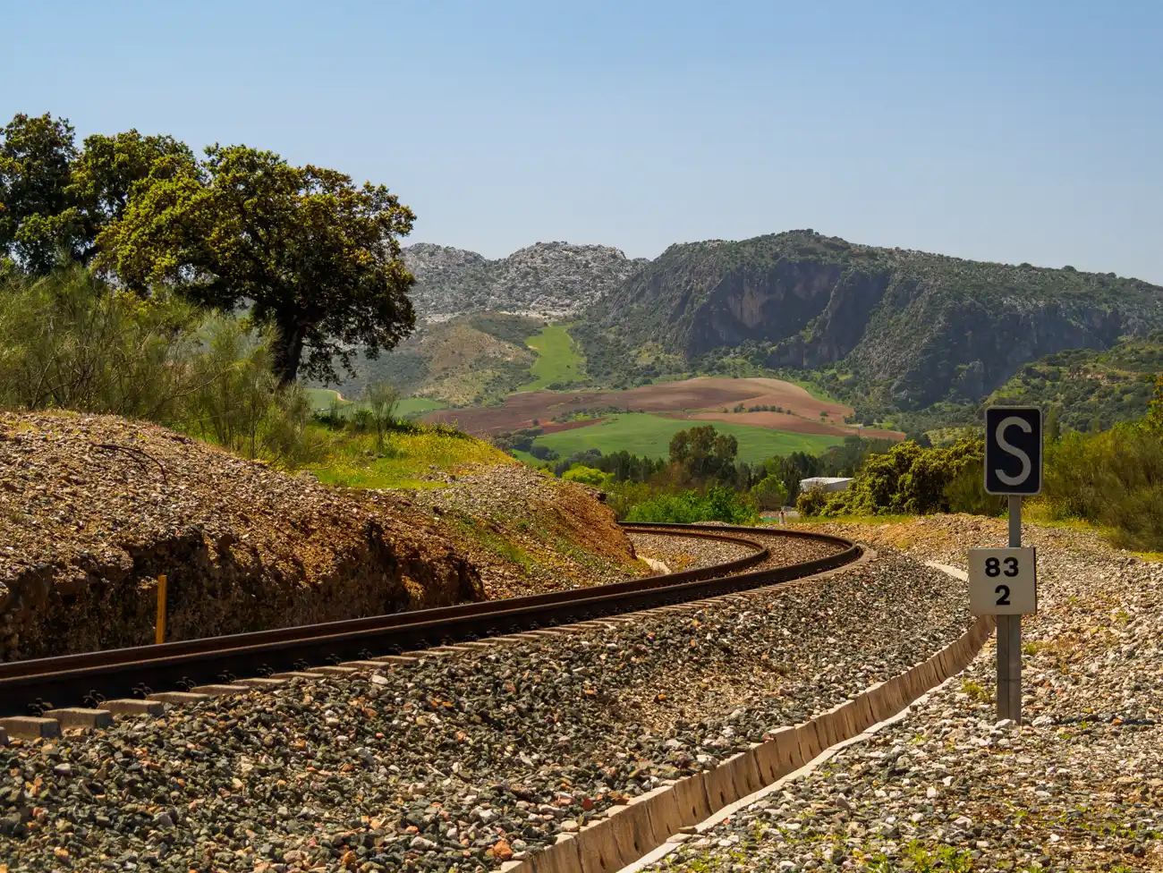 Vía férrea única que describe una curva entre desmontes de balasto, con un poste de señal y un hito kilométrico, y sierras y campos de cultivo al fondo.