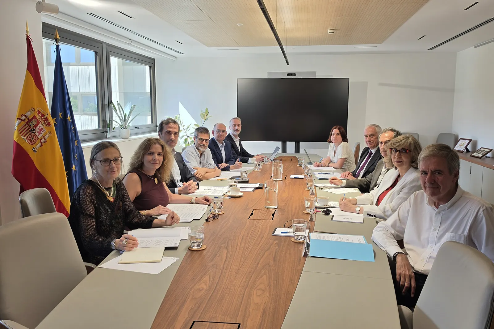 Una decena de personas sentadas alrededor de una mesa alargada de madera en una sala de reuniones con banderas de España y de la Unión Europea, carpetas y documentos sobre la mesa y una pantalla apagada al fondo.