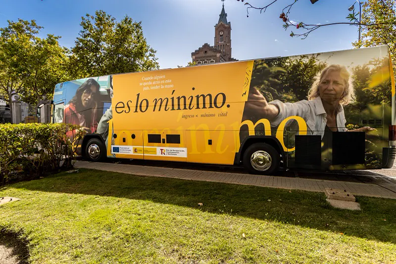Autobús amarillo rotulado con el lema «es lo mínimo. ingreso mínimo vital» y fotografías de dos personas, aparcado junto a un seto en una calle arbolada.