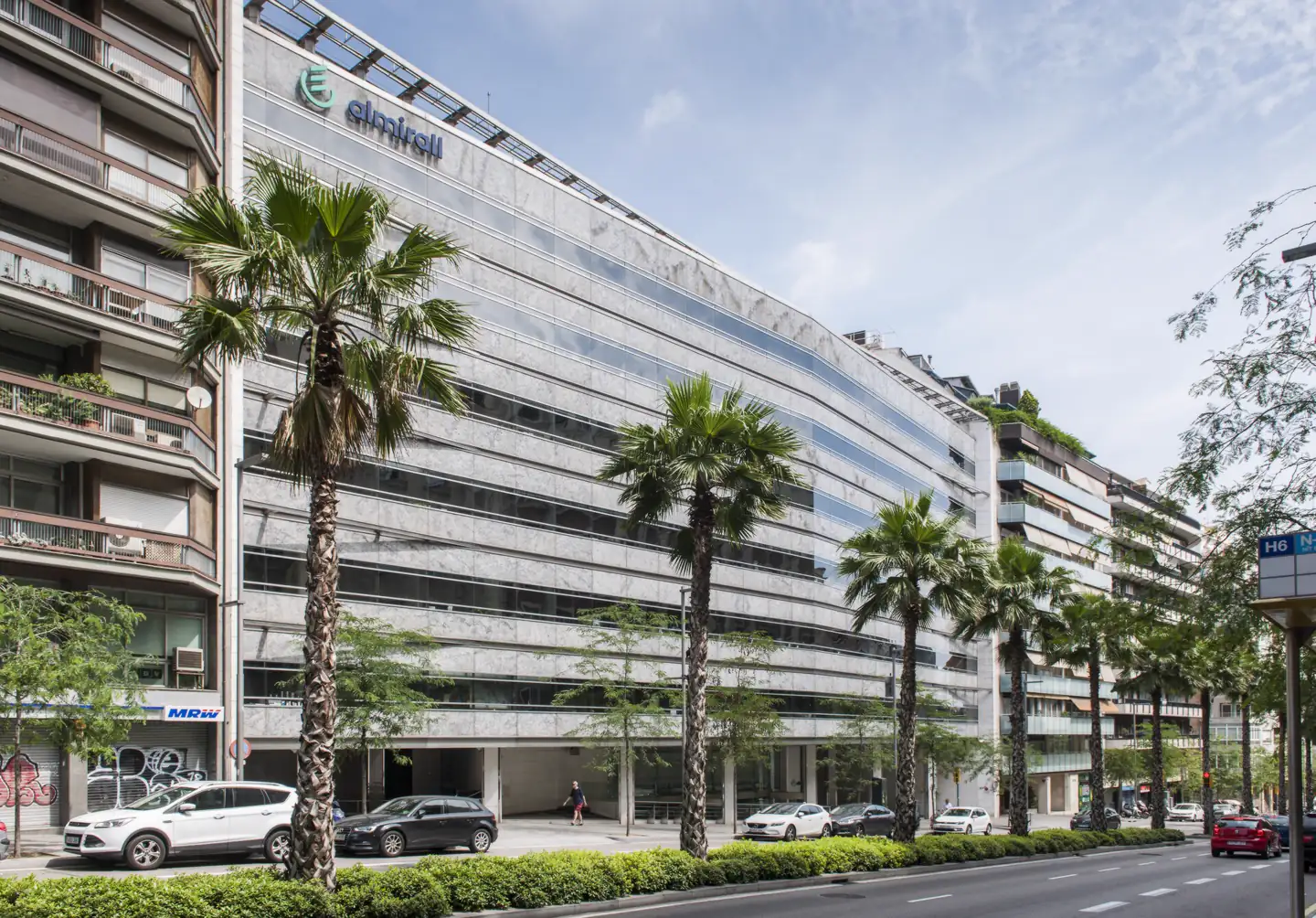 Fachada acristalada de la sede de Almirall en Barcelona, con el logotipo de la compañía en lo alto del edificio y una hilera de palmeras en la acera.