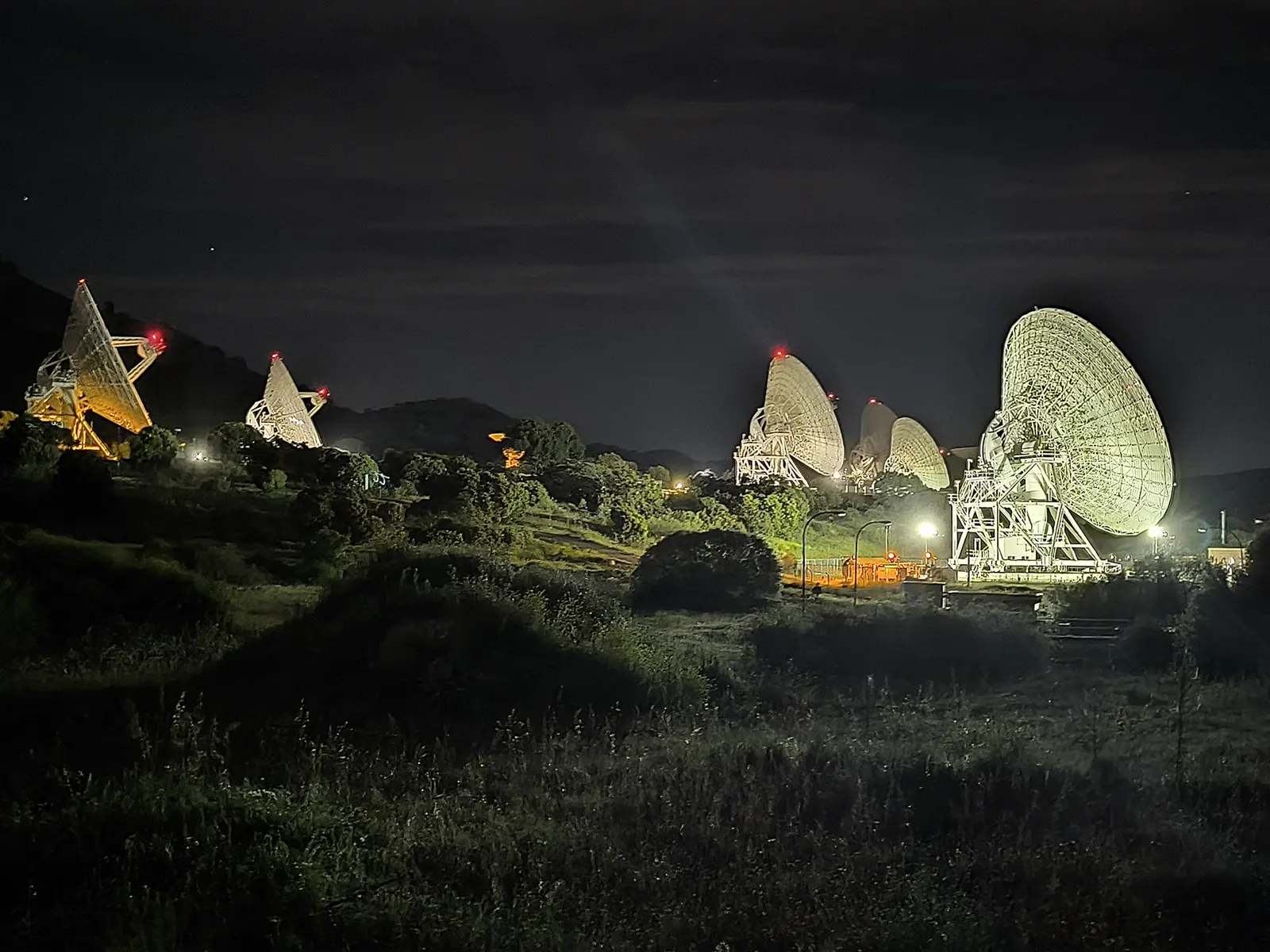 Seis grandes antenas parabólicas iluminadas de noche, apuntando en la misma dirección, sobre laderas de monte bajo y arbolado en el complejo de Robledo de Chavela.