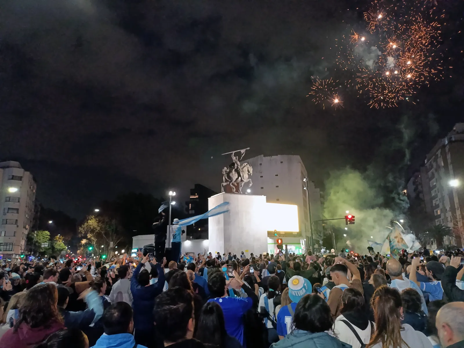 Una multitud con camisetas y banderas argentinas celebra de noche junto a un monumento ecuestre en Buenos Aires, con fuegos artificiales en el cielo