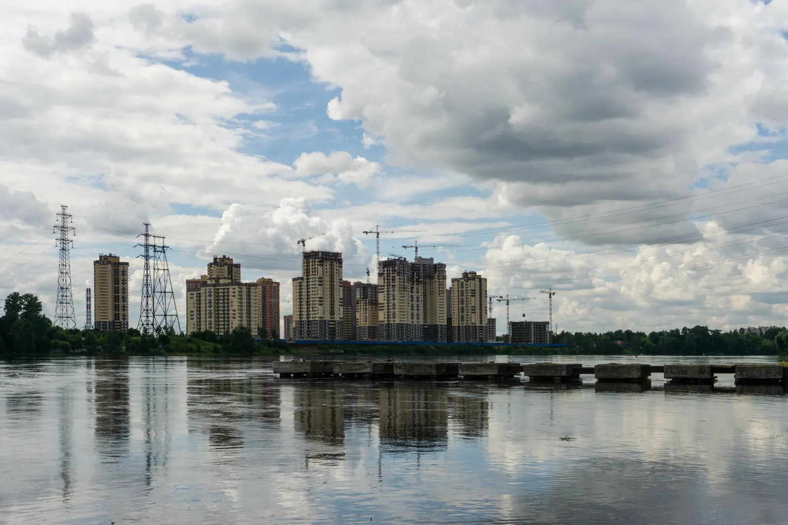 Vista del ancho río Nevá desde una orilla baja; en la ribera de enfrente, un grupo de bloques de viviendas en construcción con grúas torre y dos torres de alta tensión, bajo un cielo de nubes.