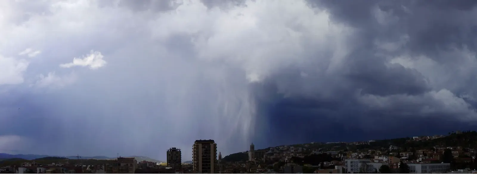 Cortina de lluvia cayendo desde una gran nube oscura de tormenta sobre los tejados y los bloques de la ciudad de Girona, con las colinas boscosas a la derecha.