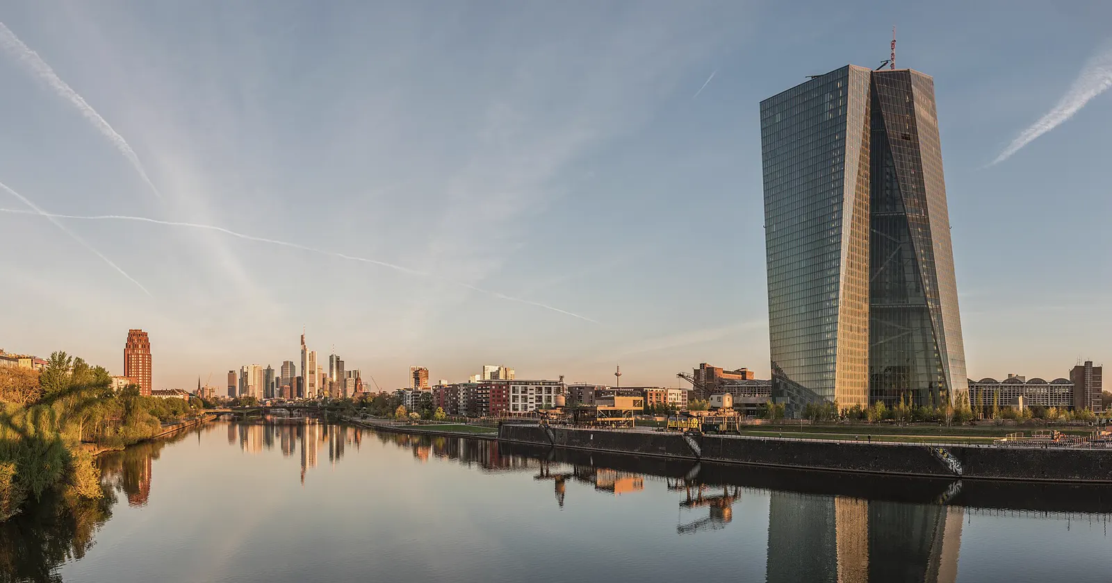 La torre de cristal del Banco Central Europeo, junto al río Meno, reflejada en el agua al amanecer, con el perfil de rascacielos de Fráncfort al fondo