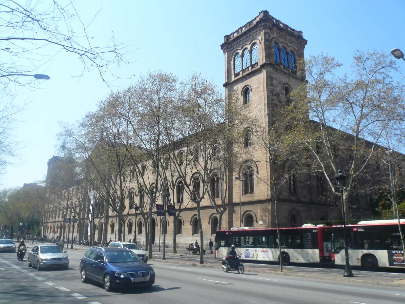 Fachada de piedra del edificio histórico de la Universidad de Barcelona, con su torre y sus ventanales de arco, vista desde la Gran Via entre plátanos sin hojas, con coches y un autobús urbano circulando delante.