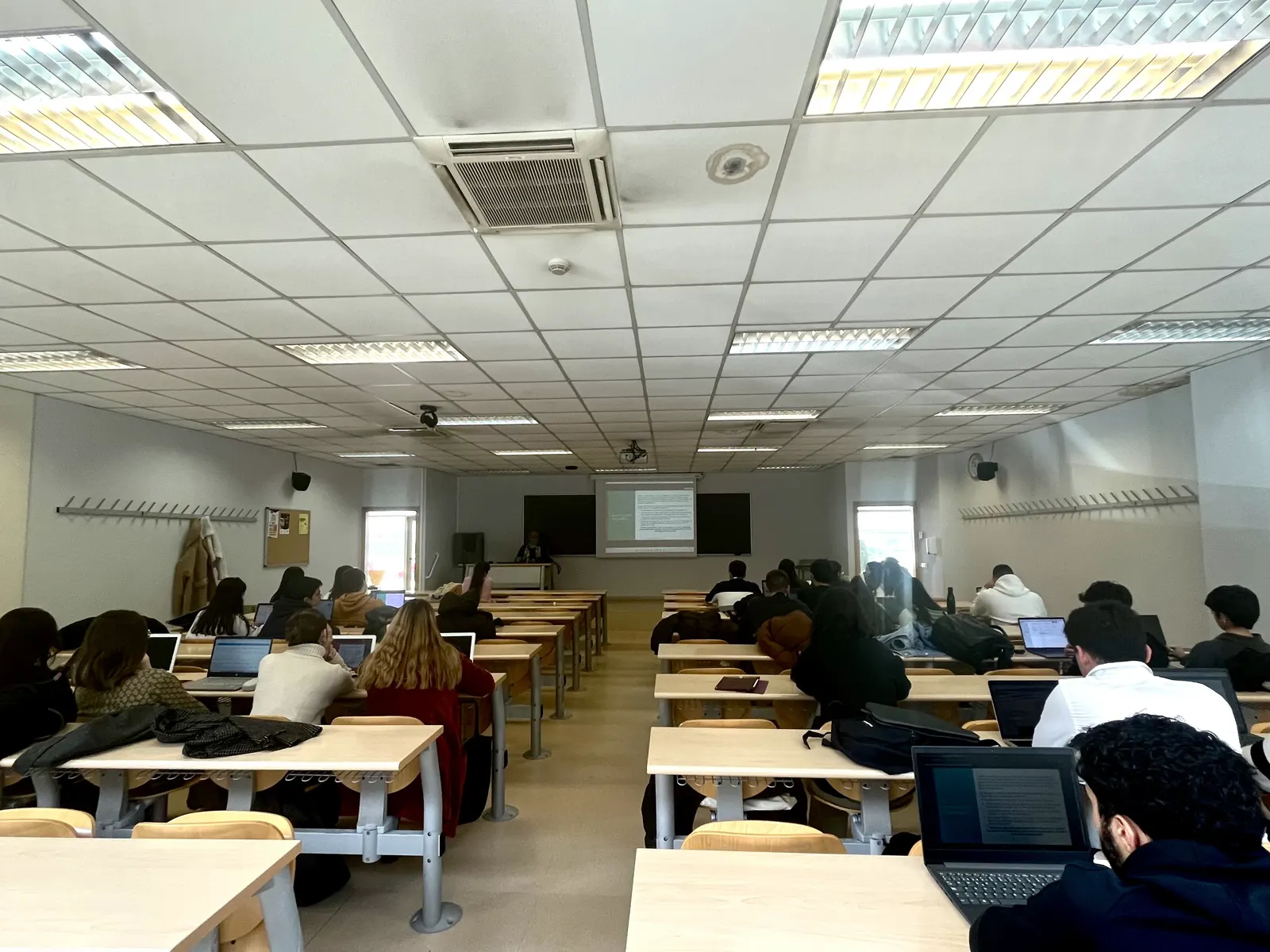 Aula universitaria vista desde el fondo: filas de pupitres con estudiantes de espaldas trabajando con portátiles mientras una profesora imparte clase junto a una pantalla proyectada.