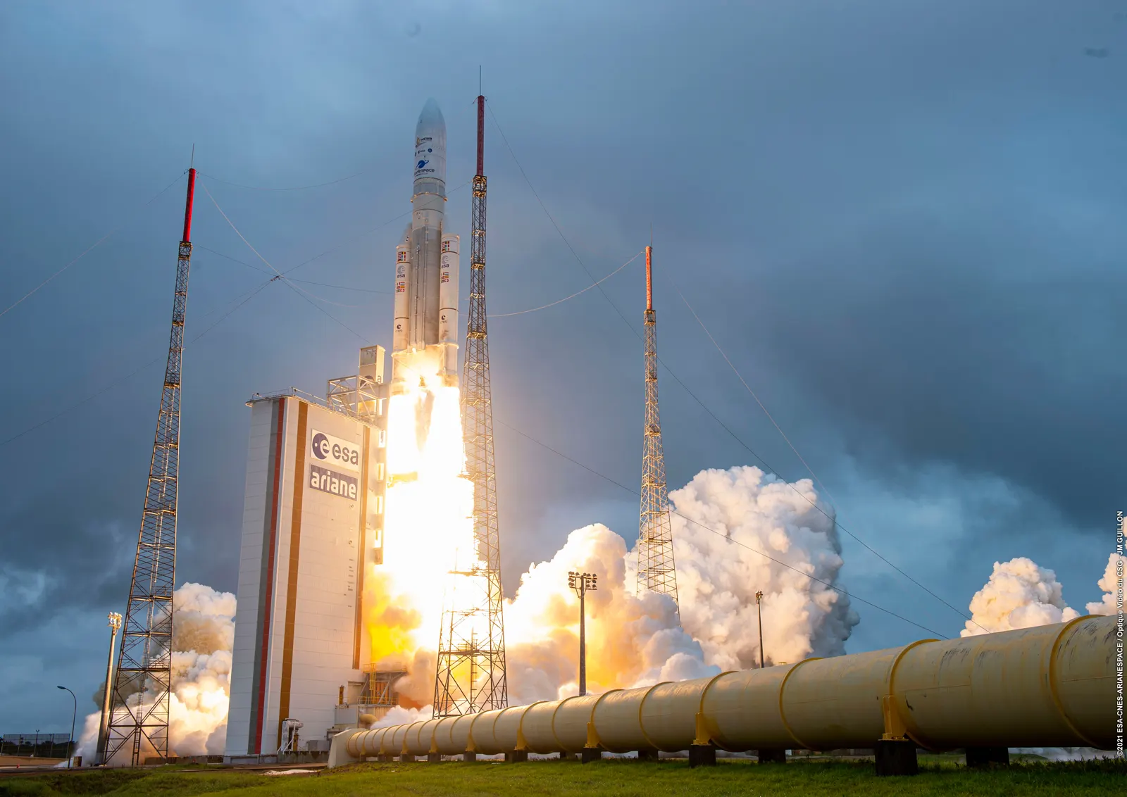 Un cohete Ariane 5 despega entre llamas y una gran nube de humo blanco desde su plataforma de lanzamiento, junto a una torre con el rótulo «esa ariane» y varios mástiles pararrayos.