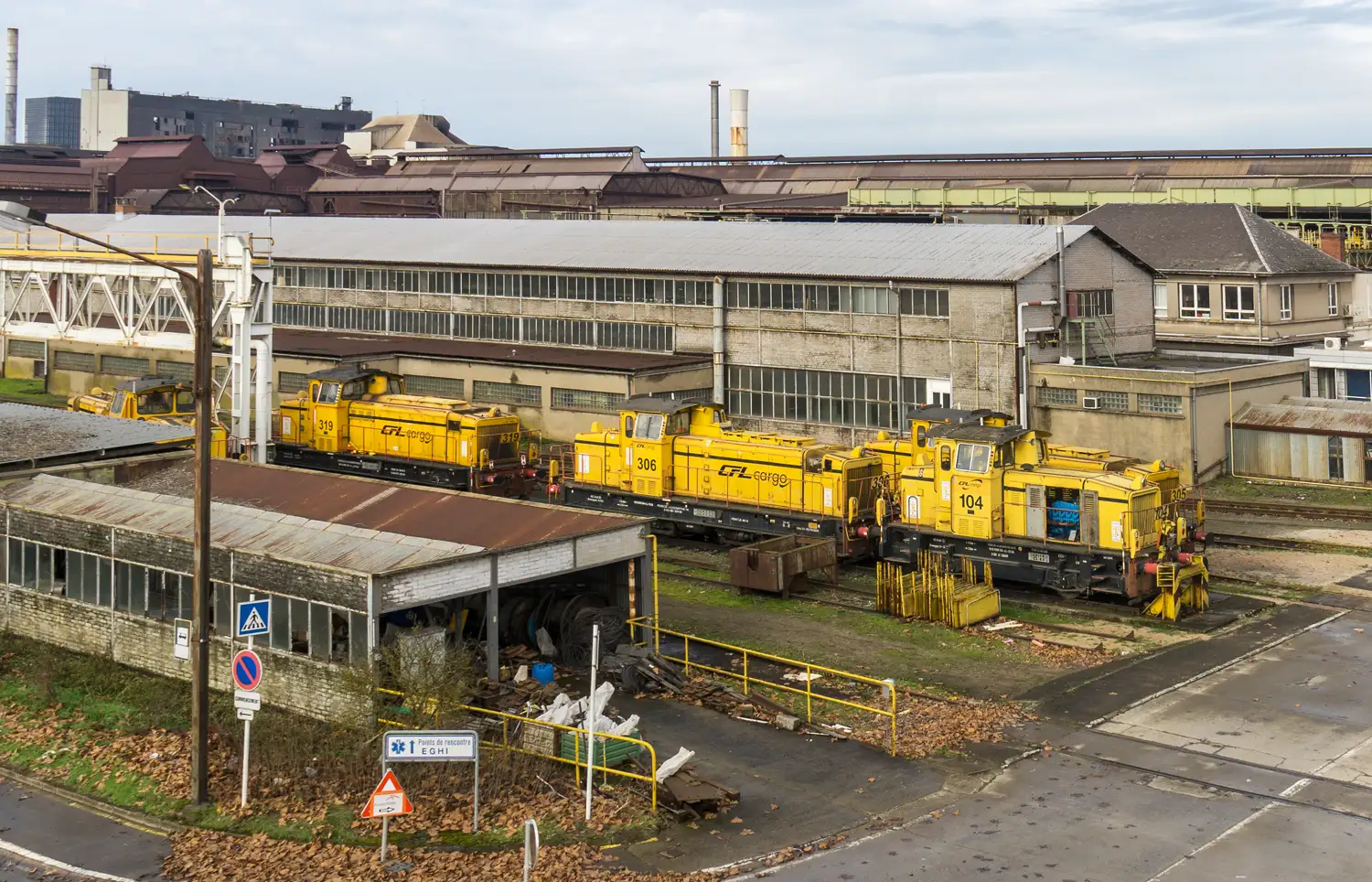 Cuatro locomotoras diésel amarillas de CFL Cargo estacionadas en las vías junto a naves industriales de la acería de Esch-Belval, en Luxemburgo, bajo un cielo gris.