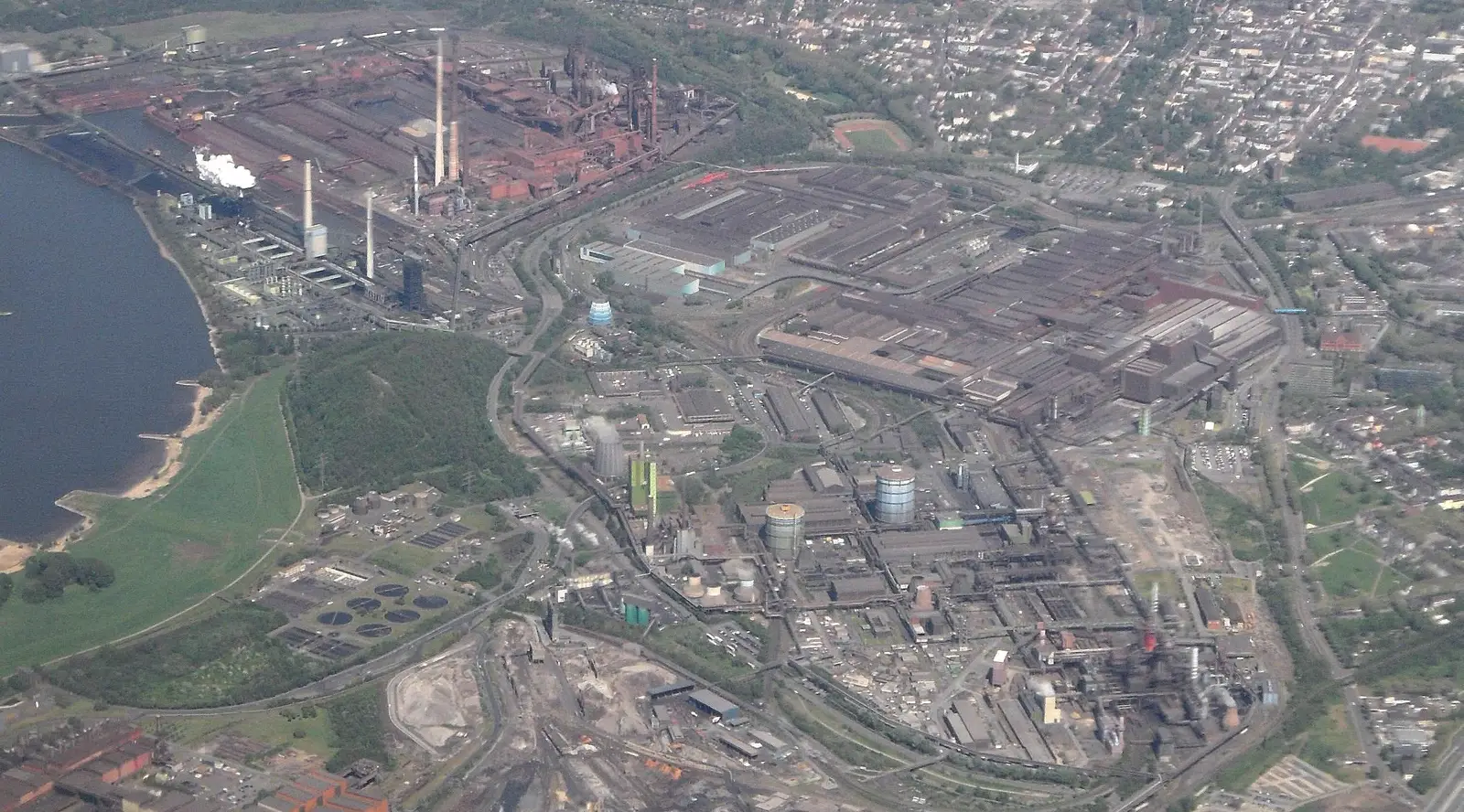Vista aérea de un extenso complejo siderúrgico junto a un río, con altos hornos, chimeneas humeantes, naves de laminación y barrios residenciales alrededor.