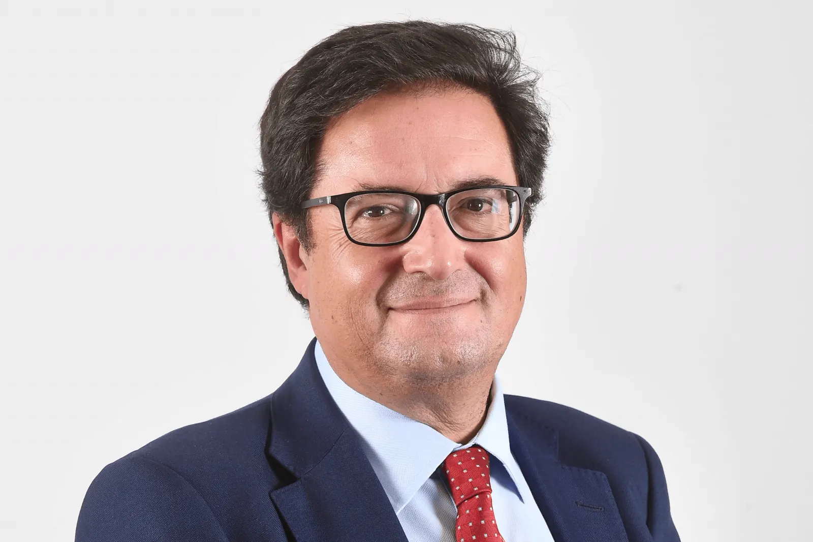 Retrato de estudio de Óscar López, ministro para la Transformación Digital y de la Función Pública, con gafas, traje azul marino y corbata roja, sobre fondo blanco.
