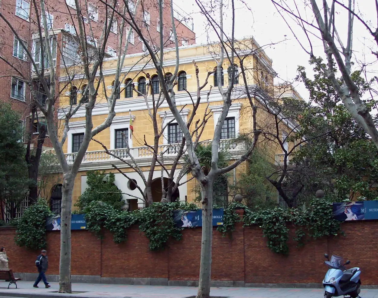 Fachada amarilla de un palacete de dos plantas tras una tapia de ladrillo y unos plátanos sin hojas, con carteles del Museo Sorolla en la valla.