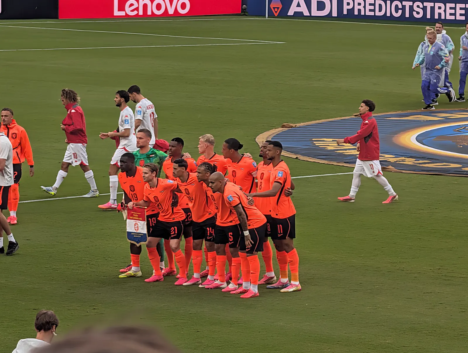 Los once titulares de Países Bajos, con camiseta naranja y en dos filas, posan sobre el césped antes de un partido; al fondo pasan suplentes con chándal rojo y operarios con la bandera de la FIFA