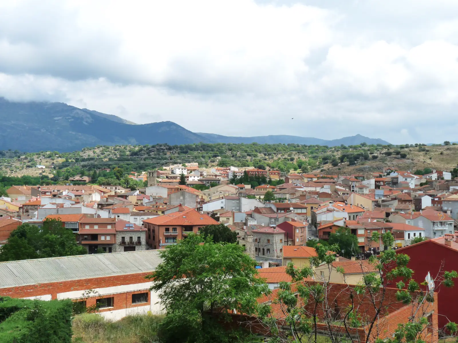 Vista del casco urbano de Burgohondo (Ávila), con tejados de teja roja escalonados en la ladera y las estribaciones de la sierra al fondo bajo un cielo nuboso.