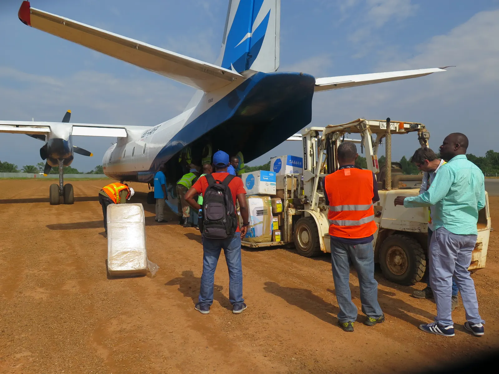 Operarios y personal sanitario descargan cajas de material médico de un avión de hélice en una pista de tierra roja, con una carretilla elevadora al lado