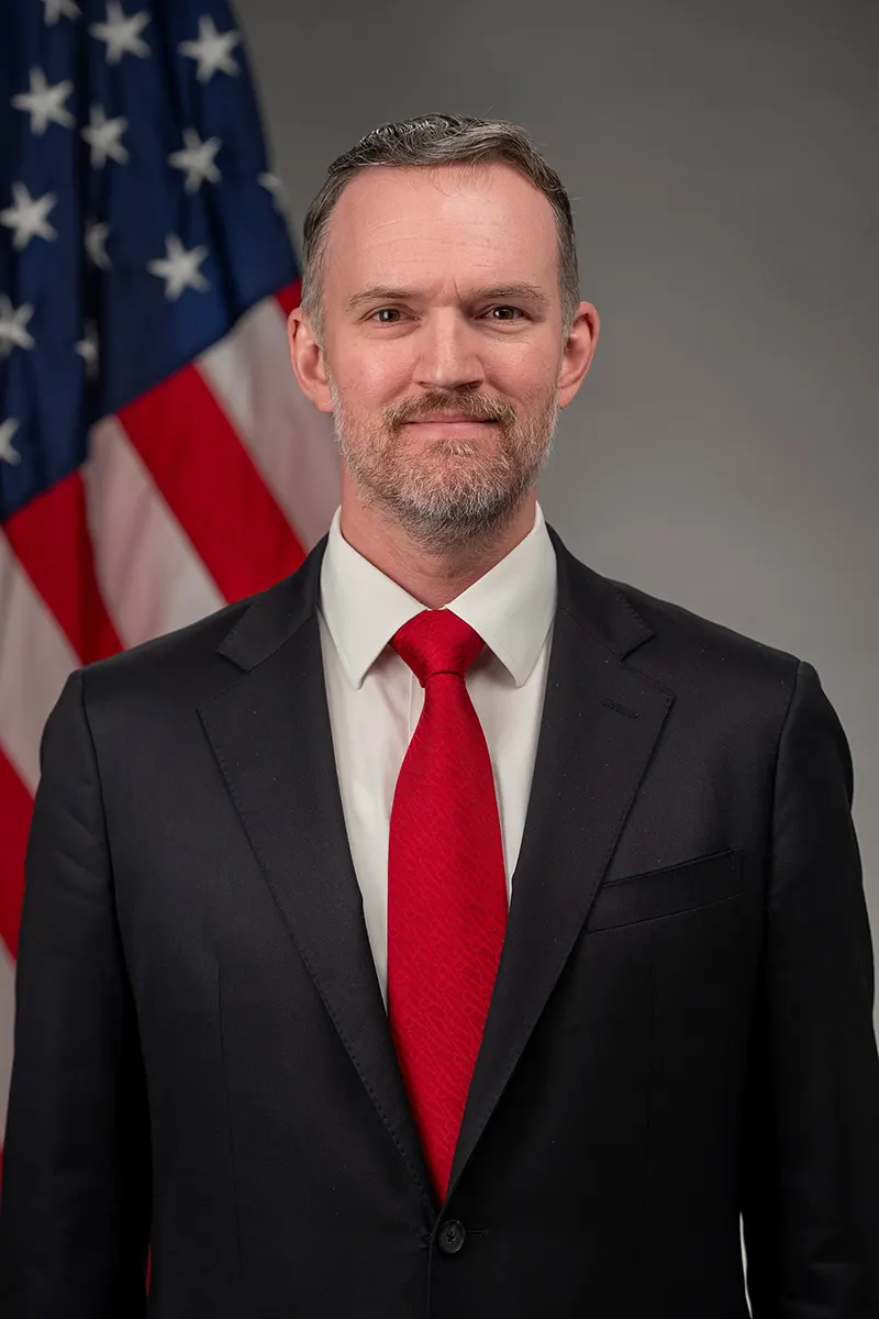 Retrato oficial de Jamieson Greer, representante comercial de Estados Unidos, con traje y corbata ante la bandera estadounidense.
