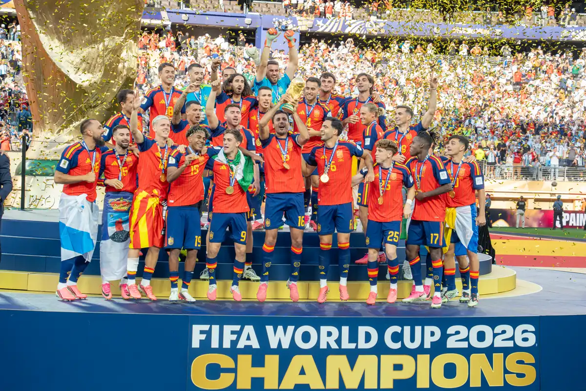 La selección española celebra en el podio con la Copa del Mundo en alto bajo una lluvia de confeti dorado, ante un cartel que dice FIFA World Cup 2026 Champions