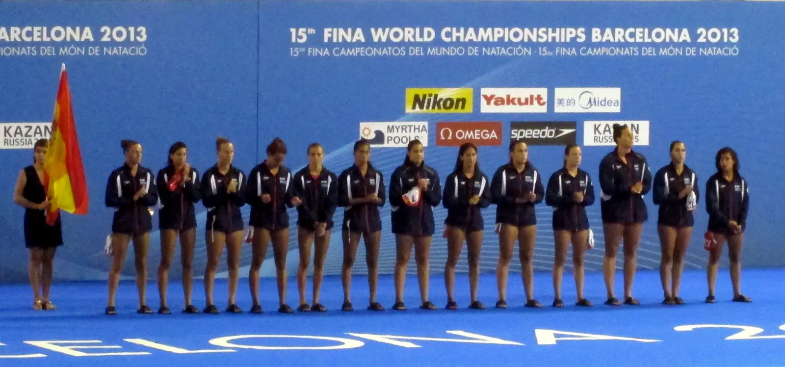 Las jugadoras de la selección española de waterpolo, en fila y con chándal azul marino, escuchan firmes junto a una portabanderas con la bandera de España ante el panel del Mundial de Barcelona 2013
