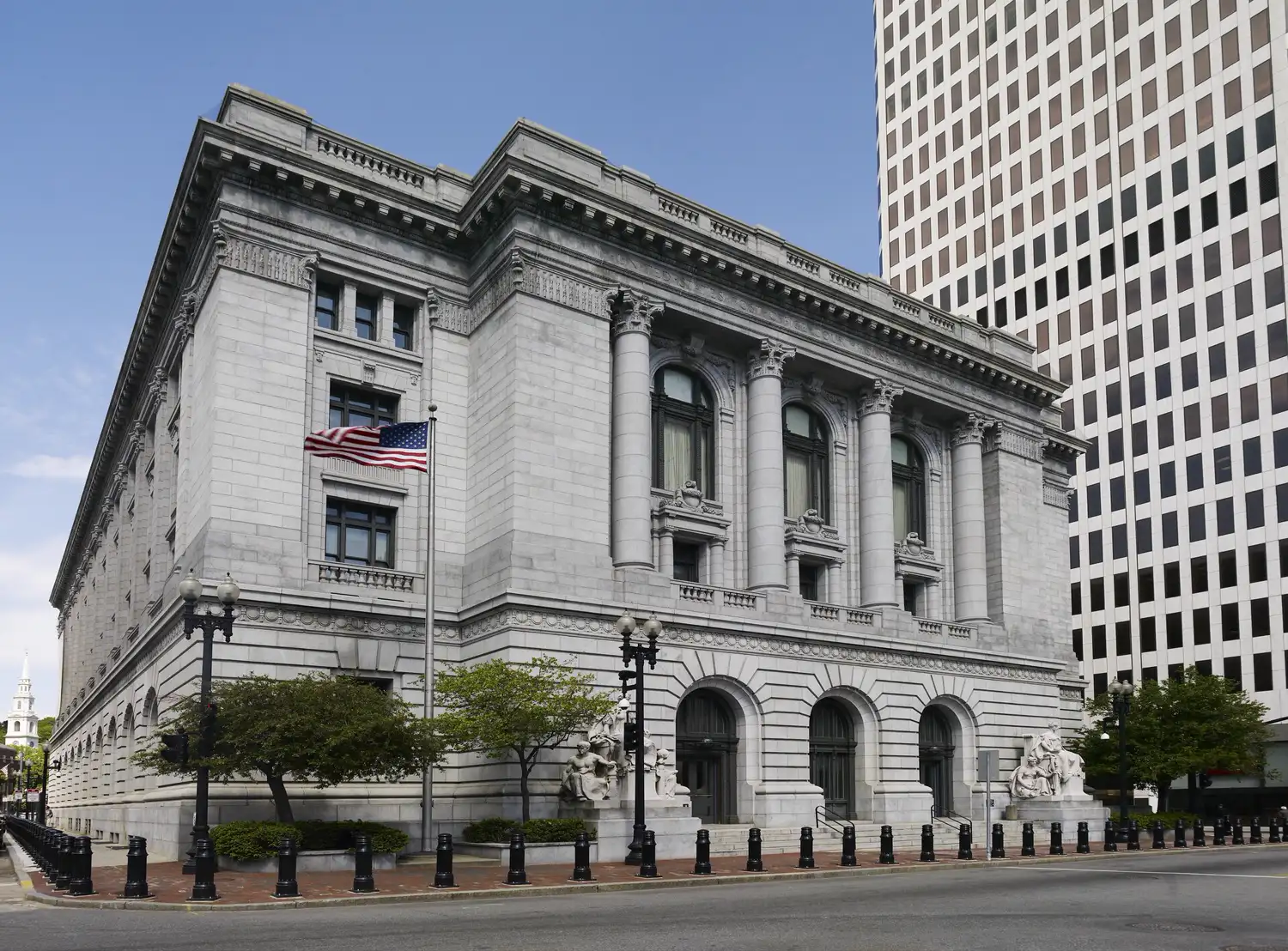 Fachada de piedra clara del edificio federal y juzgado de Providence, en Rhode Island, de estilo Beaux-Arts, con columnas, esculturas junto a la entrada y una bandera de Estados Unidos en un mástil.