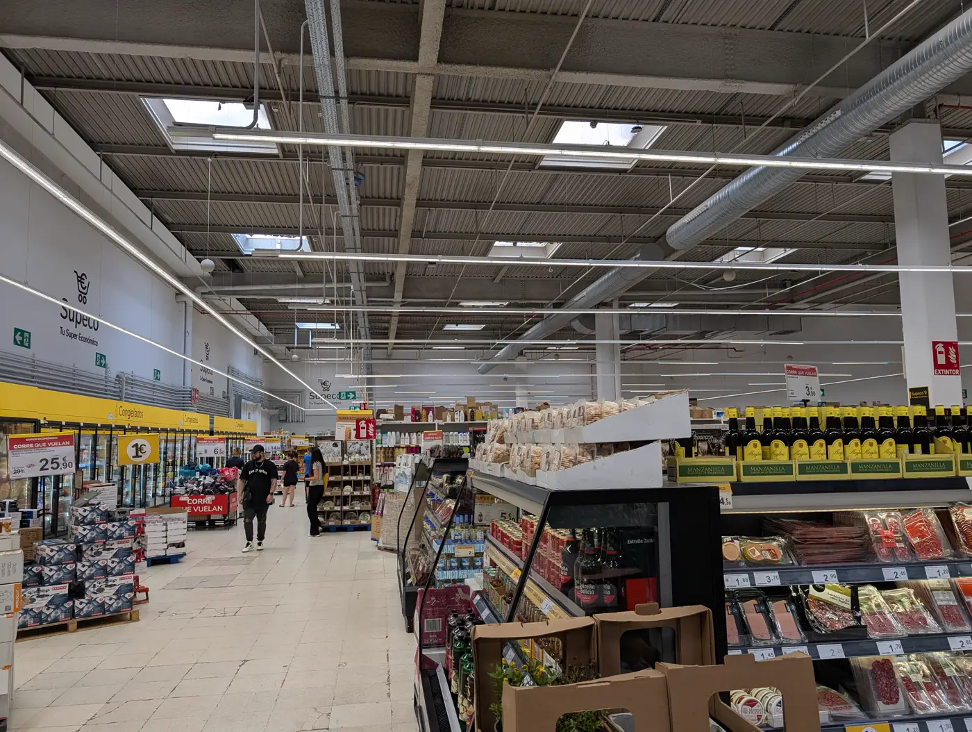 Pasillo central de un supermercado con carteles de ofertas y precios en euros a ambos lados y varios clientes al fondo