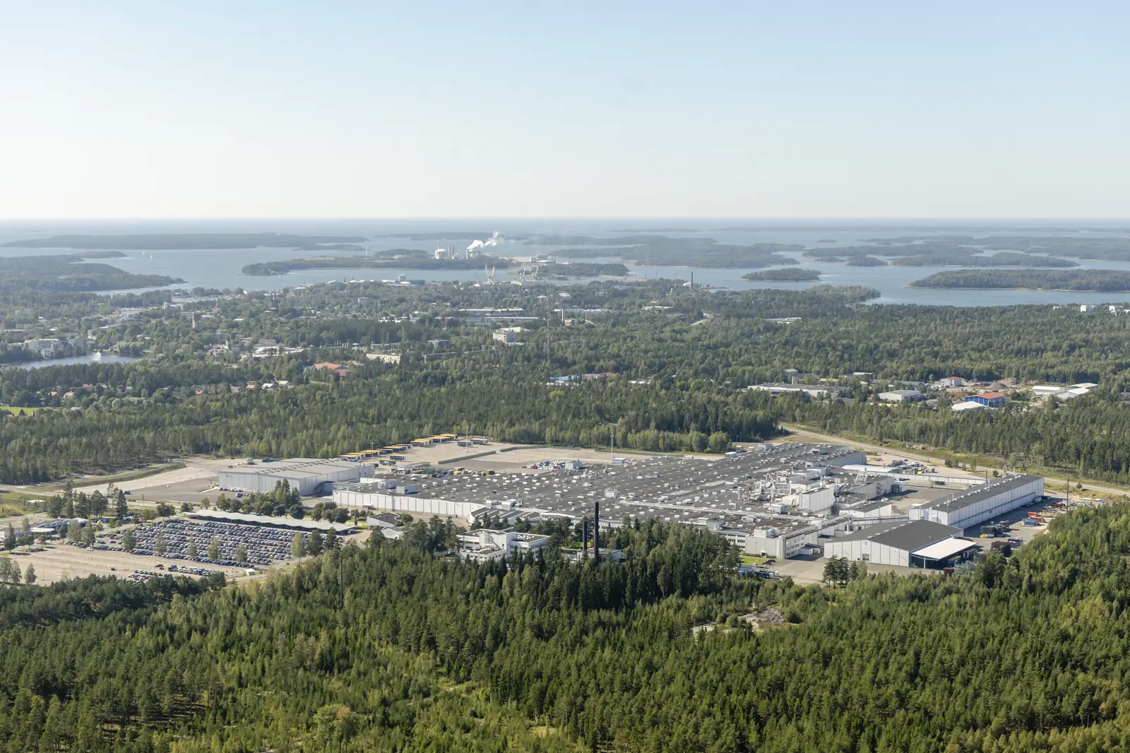 Vista aérea de la fábrica de automóviles de Valmet Automotive en Uusikaupunki: naves industriales blancas de cubierta gris rodeadas de bosque, con un aparcamiento lleno de coches y, al fondo, la ciudad y el archipiélago finlandés.