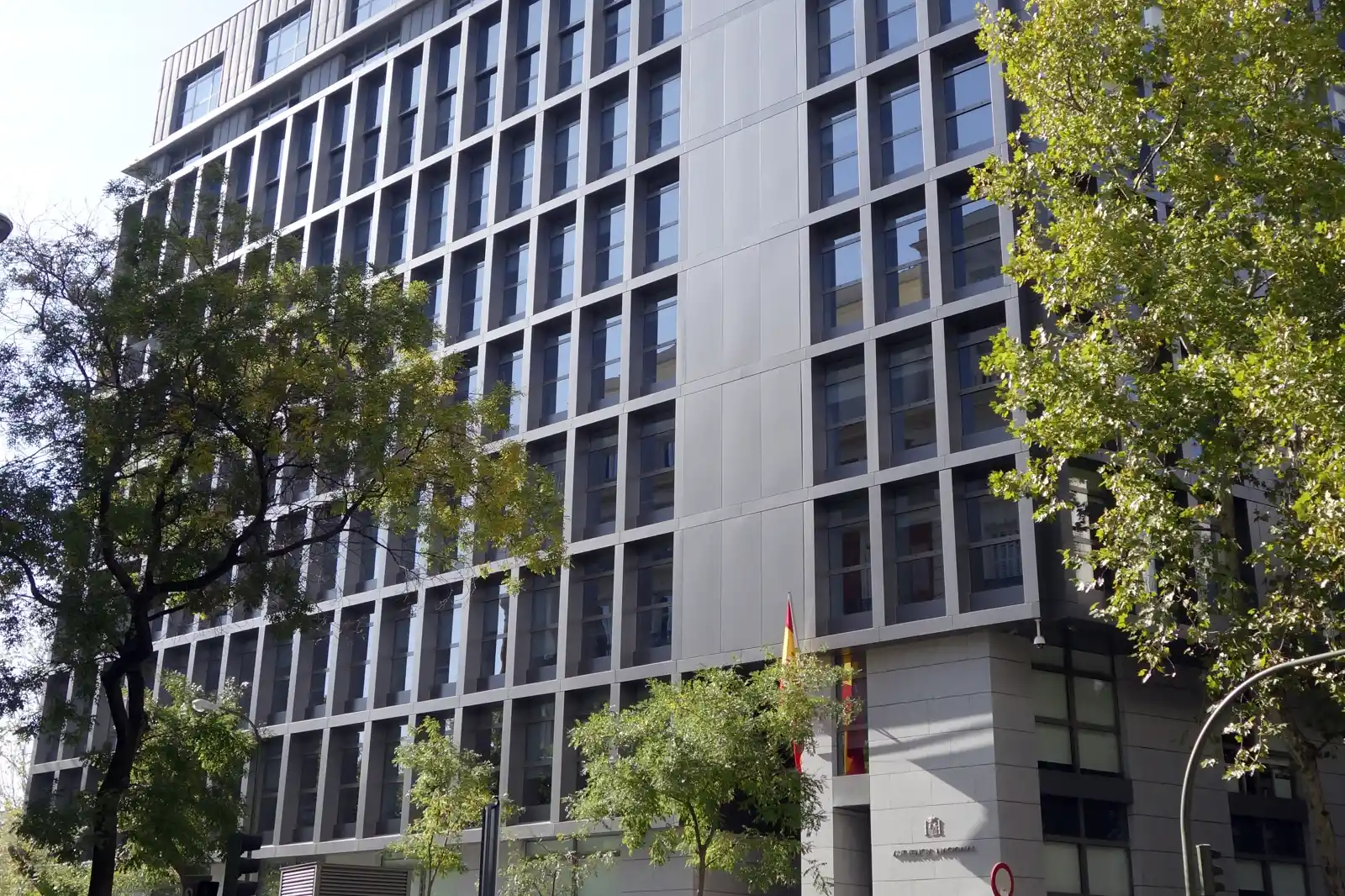 Fachada acristalada del edificio de la Audiencia Nacional en Madrid, con la bandera de España sobre la entrada y árboles en la acera.