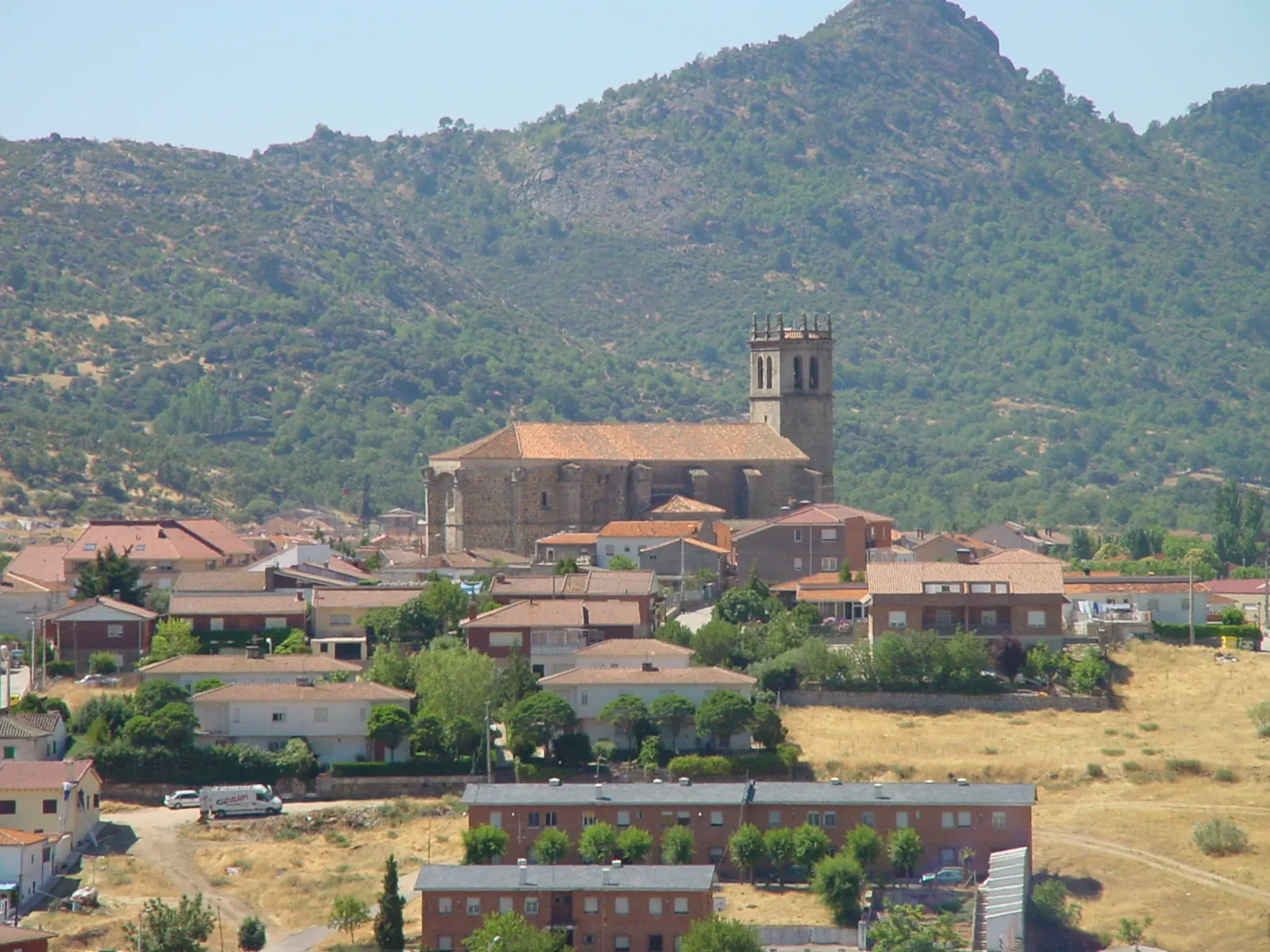 Vista de Robledo de Chavela: casas de teja roja y la iglesia de la Asunción, con su torre almenada, al pie de una ladera de monte bajo y arbolado.