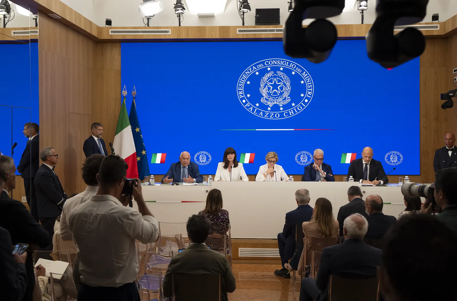 Cinco miembros del Gobierno italiano sentados tras una mesa larga en la sala de prensa de Palazzo Chigi, ante el emblema de la Presidencia del Consejo de Ministros, rodeados de periodistas y cámaras.