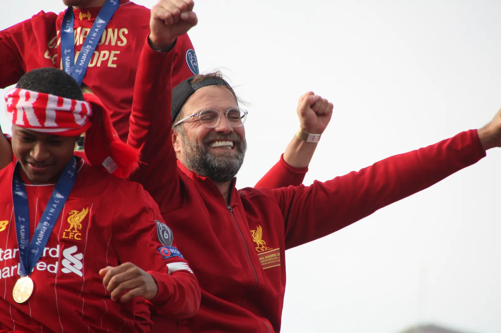 Jürgen Klopp, con chándal rojo del Liverpool y gafas, sonríe con los brazos en alto rodeado de jugadores durante un desfile en autobús descubierto