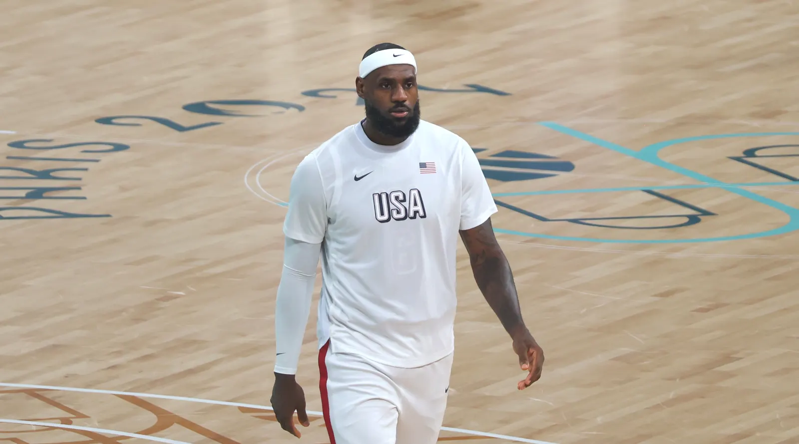 LeBron James, con camiseta blanca de calentamiento de la selección de Estados Unidos y cinta en la cabeza, camina por una pista de baloncesto con la pintura de París 2024 en el suelo