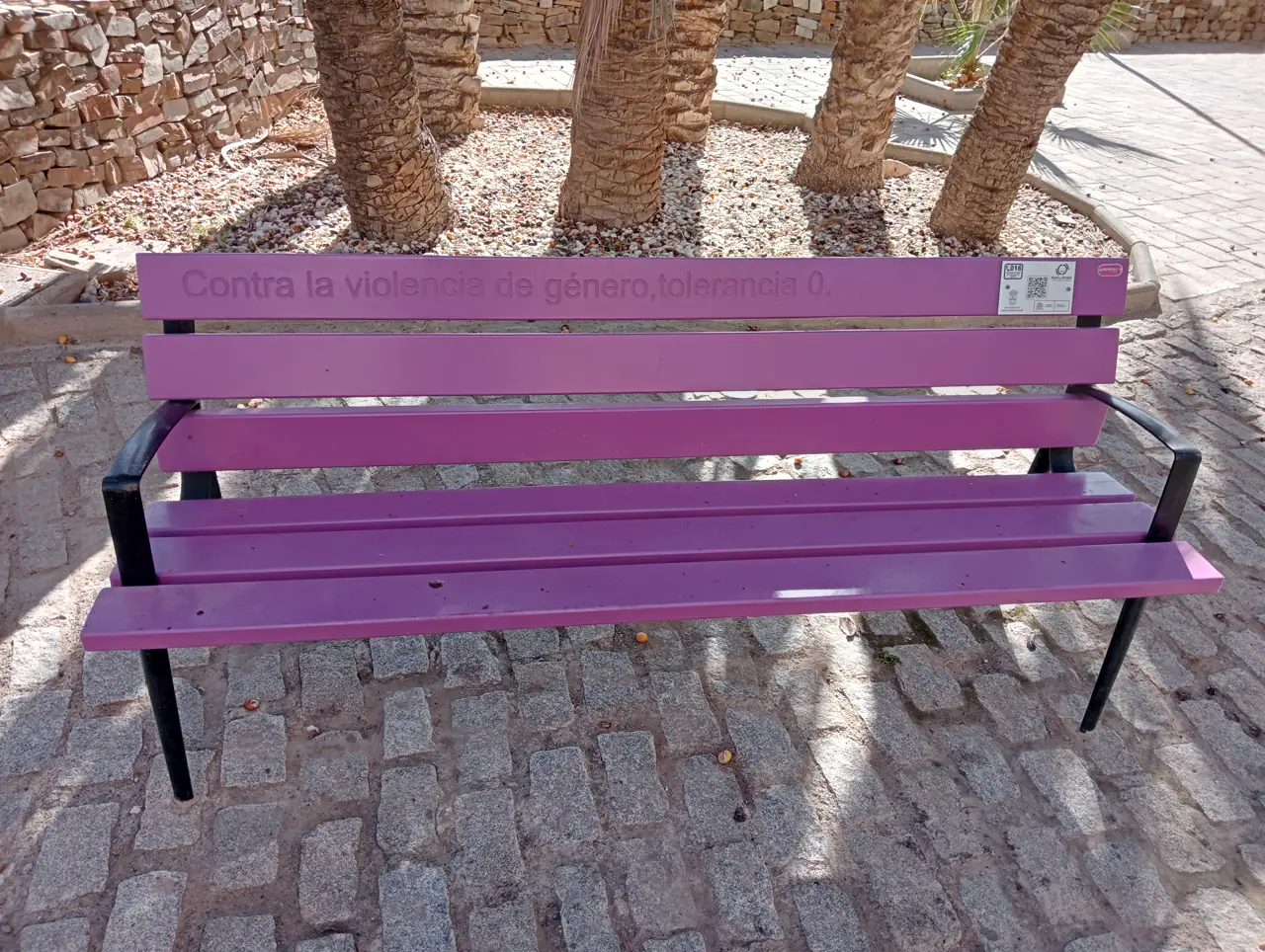 Banco público pintado de morado con la frase «Contra la violencia de género, tolerancia 0» grabada en el respaldo, en una plaza empedrada con palmeras