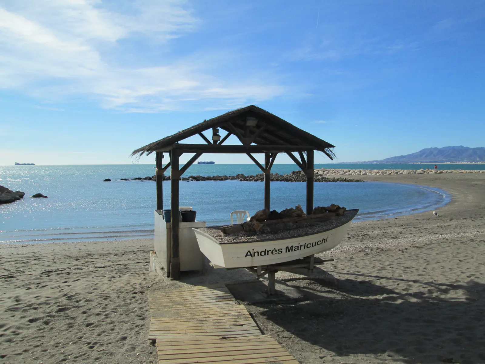 Playa de Pedregalejo, en Málaga: una barca de madera con techado de paja sobre la arena, junto a un espigón de piedra y el mar en calma, con la bahía y las montañas al fondo.