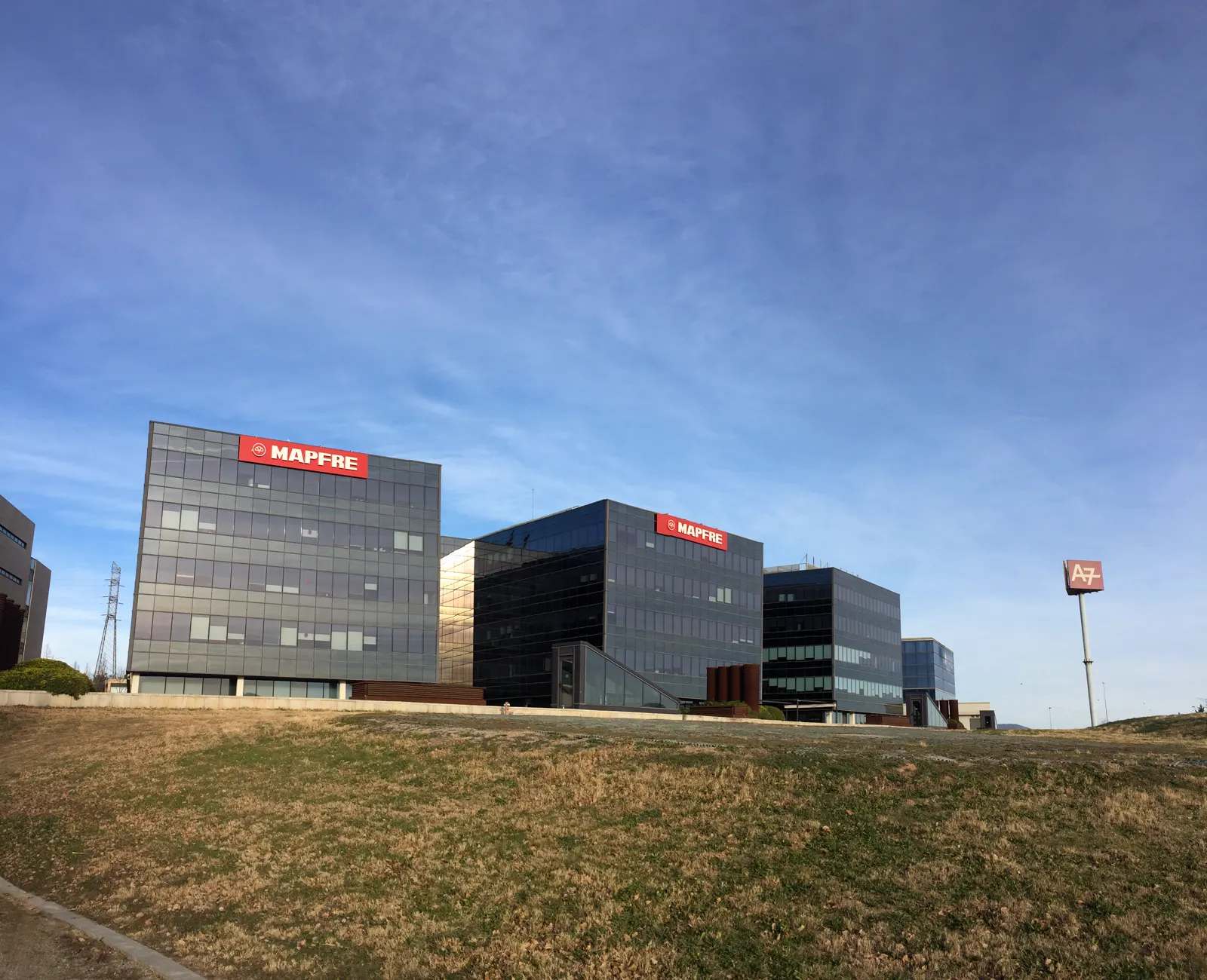 Edificios acristalados con el logotipo rojo de Mapfre en la fachada, bajo un cielo azul, junto a un poste con el rótulo A7.