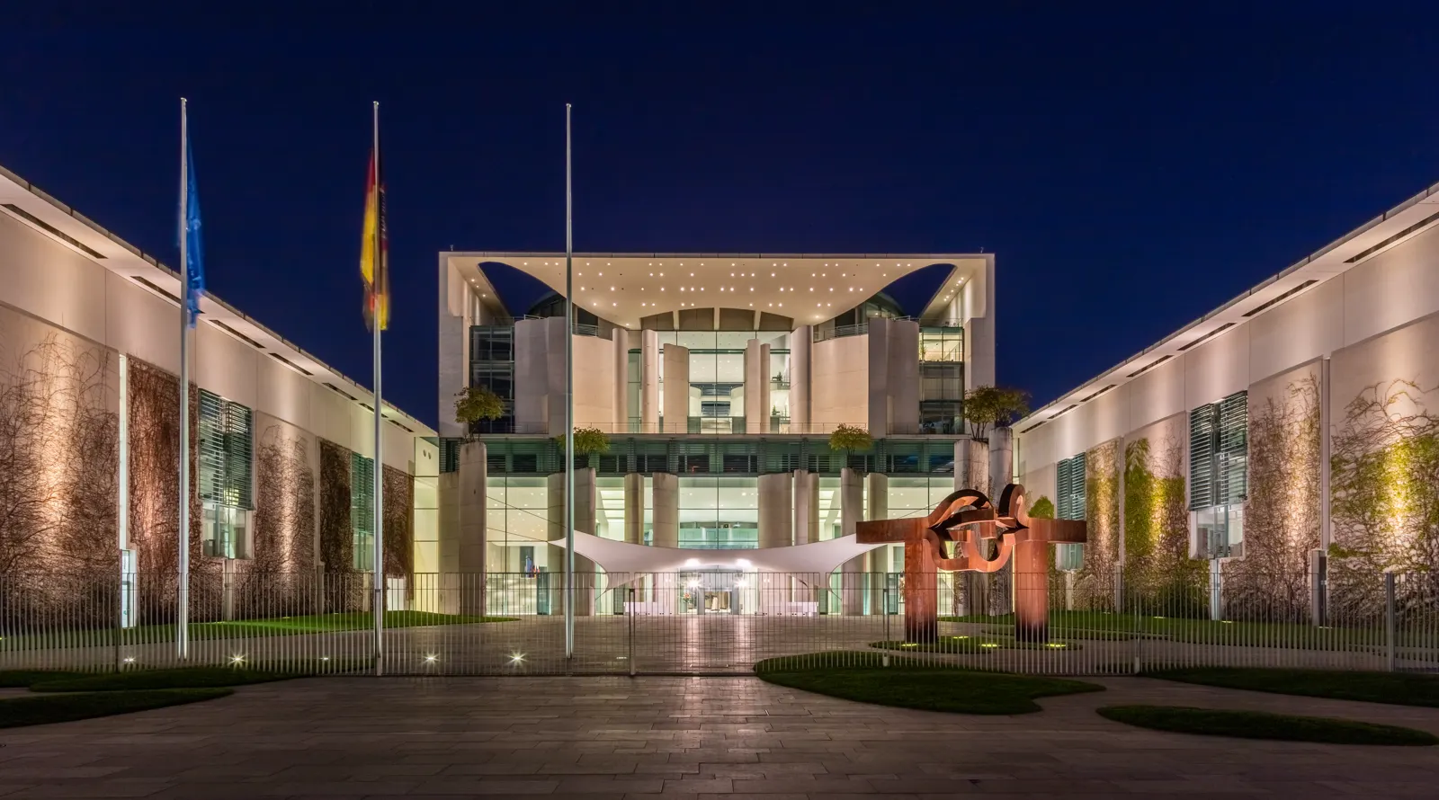 Fachada principal de la Cancillería Federal alemana en Berlín, iluminada de noche, con las banderas de la Unión Europea y de Alemania en el patio de acceso y una escultura de acero rojizo a la derecha.