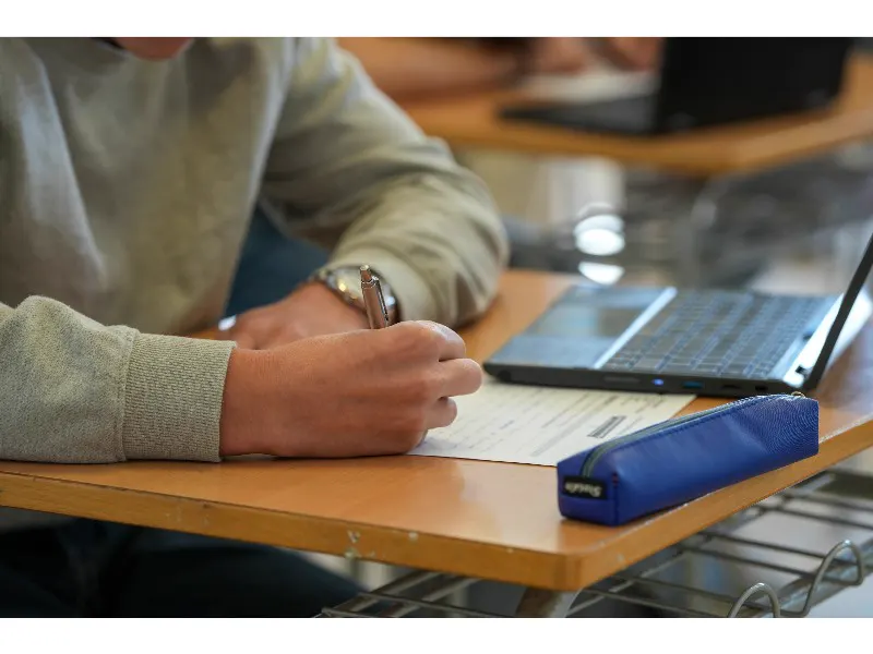 Primer plano de las manos de un estudiante rellenando a bolígrafo un cuestionario sobre un pupitre, junto a un ordenador portátil y un estuche azul