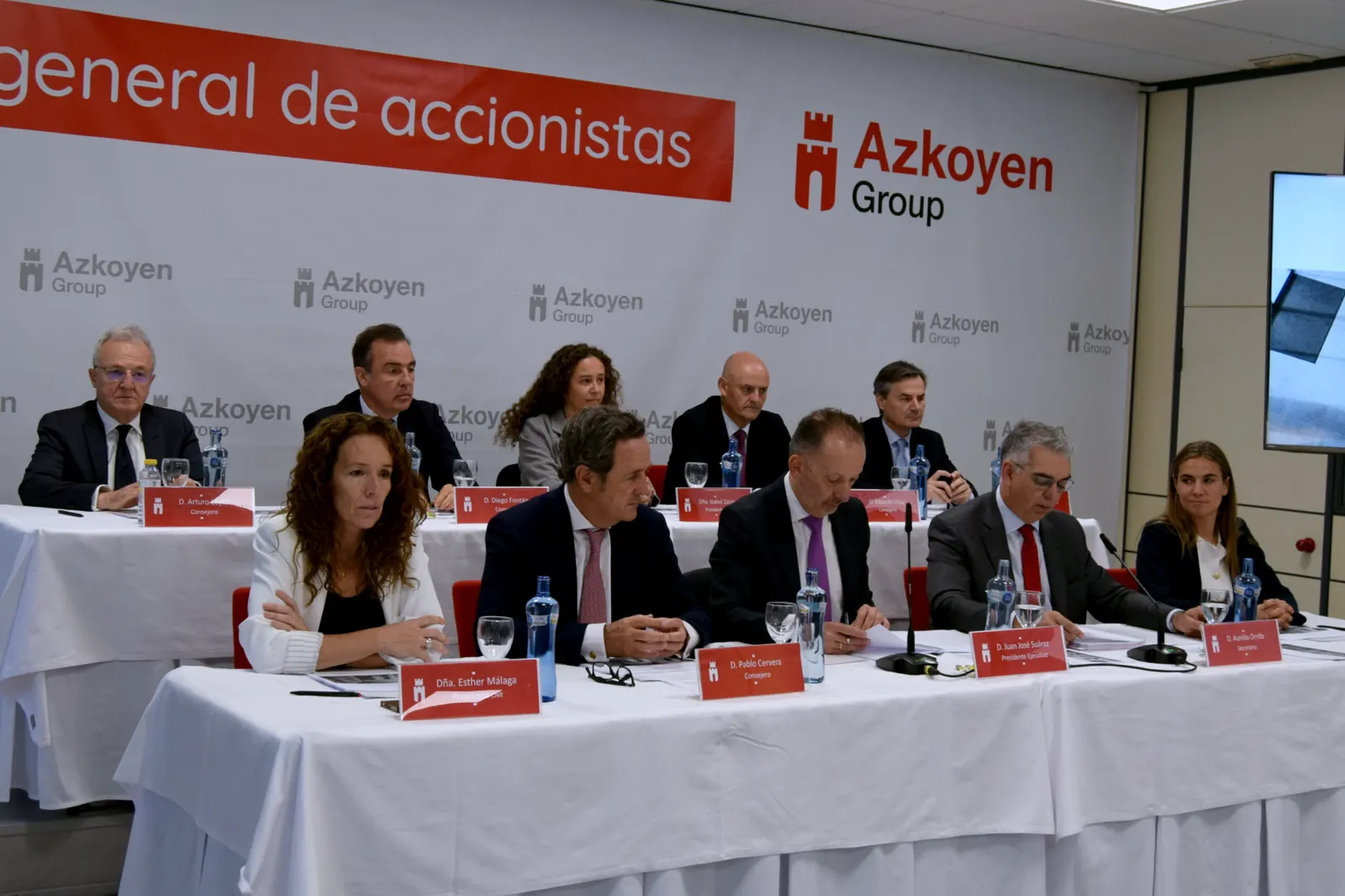 Once directivos y consejeros sentados tras una mesa alargada con mantel blanco, con carteles de nombre rojos, ante un panel con el logotipo de Azkoyen Group y el rótulo «general de accionistas».