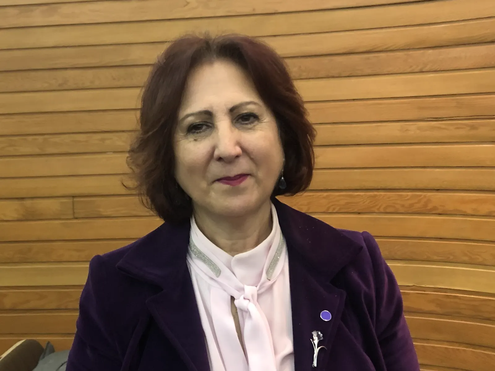 Teresa Peramato, sentada ante una pared de listones de madera, con americana morada y camisa rosa