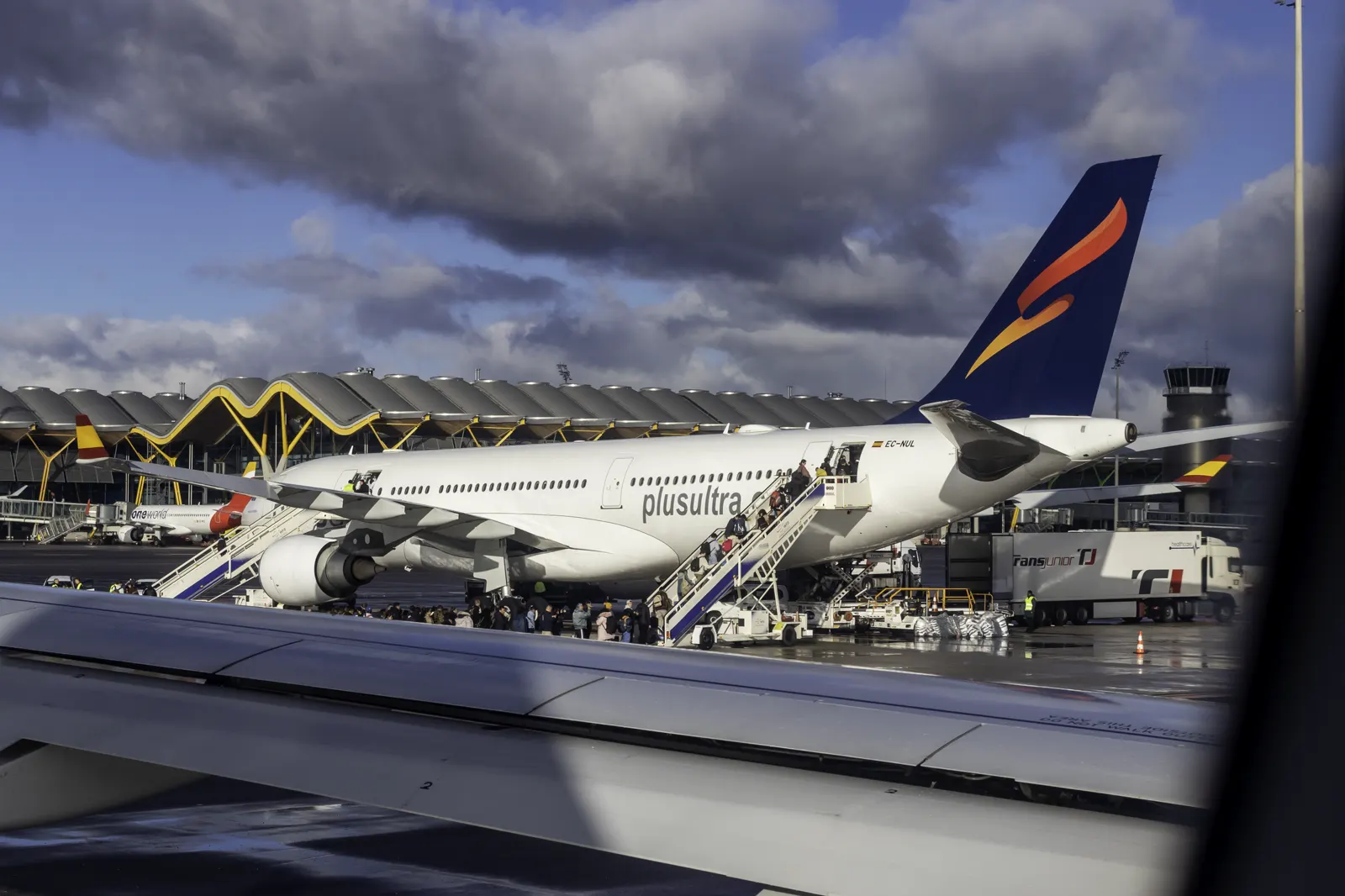 Airbus A330 de Plus Ultra Líneas Aéreas estacionado en la pista del aeropuerto Madrid-Barajas, con pasajeros subiendo por una escalerilla y la cubierta ondulada de la terminal T4 al fondo.