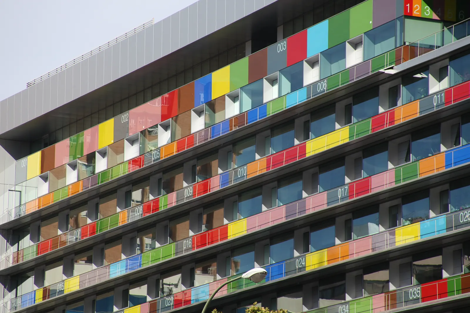 Fachada del edificio del Instituto Nacional de Estadística en Madrid, con ventanales y barandillas de vidrio de colores y los pisos numerados.