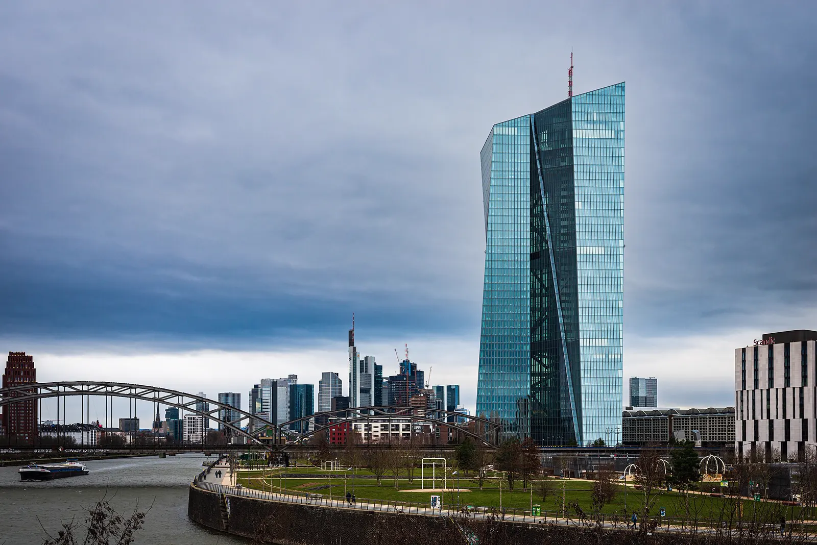 La torre de cristal del Banco Central Europeo en Fráncfort bajo un cielo nublado, con el río Meno, un puente y el perfil de rascacielos de la ciudad en segundo plano