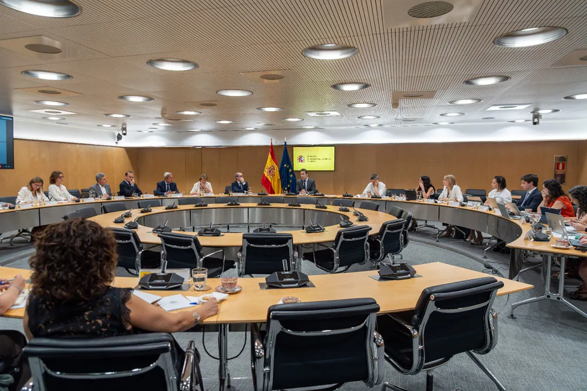 Una veintena de personas sentadas alrededor de una gran mesa ovalada en una sala de reuniones; al fondo, las banderas de España y de la Unión Europea junto al logotipo del Ministerio de Economía, Comercio y Empresa.