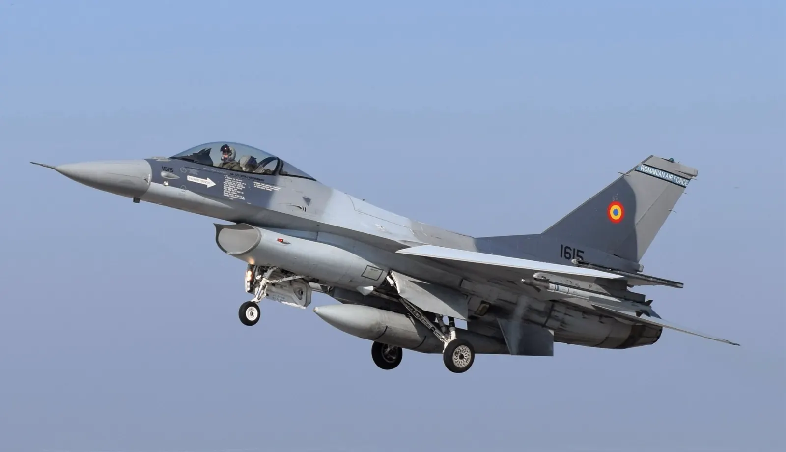 Caza F-16 de las Fuerzas Aéreas Rumanas con el tren de aterrizaje desplegado y misiles bajo las alas, volando sobre un cielo azul; en la deriva se lee «Romanian Air Force» junto a la escarapela rumana.