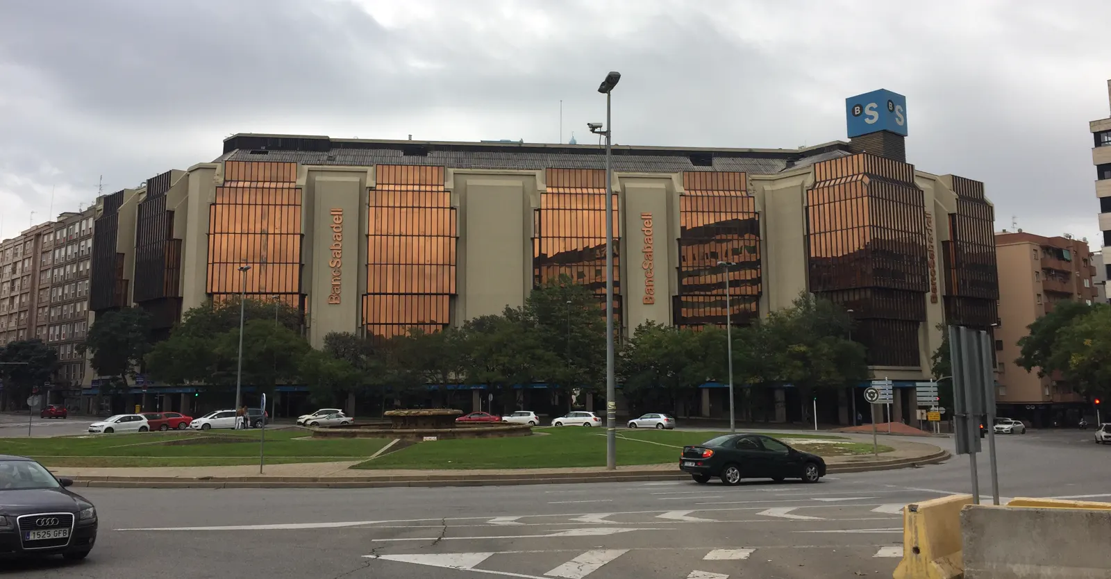 Edificio de oficinas de fachada acristalada anaranjada con el rótulo BancSabadell, junto a una rotonda con tráfico, bajo cielo nublado.