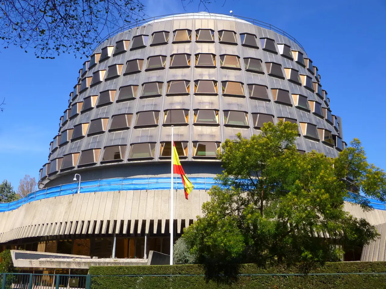 Edificio circular del Tribunal Constitucional de España, en Madrid, con su fachada de ventanales oscuros inclinados, la bandera española en un mástil ante la entrada, árboles a la derecha y el rótulo «Tribunal Constitucional» grabado en un muro bajo, abajo a la izquierda.