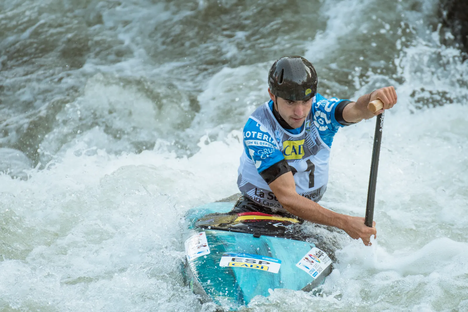 El piragüista Miquel Travé, con casco y peto de la selección española, rema entre espuma en aguas bravas a bordo de una canoa individual, con la pala de una sola hoja apoyada en el agua.