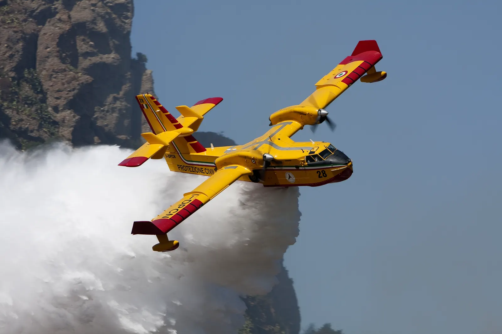 Avión anfibio Canadair CL-415 amarillo y rojo de la Protección Civil italiana, con el número 28 en el morro, soltando una gran descarga de agua sobre una ladera rocosa.