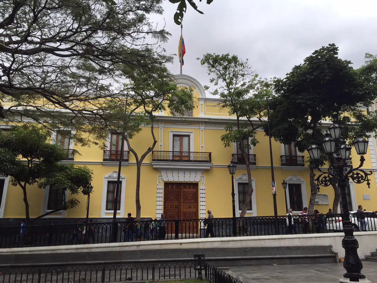 Fachada amarilla de dos plantas de la Casa Amarilla de Caracas, con balcones de hierro, portón de madera y la bandera de Venezuela ondeando sobre el frontón central; delante, árboles, farolas y viandantes tras una verja.
