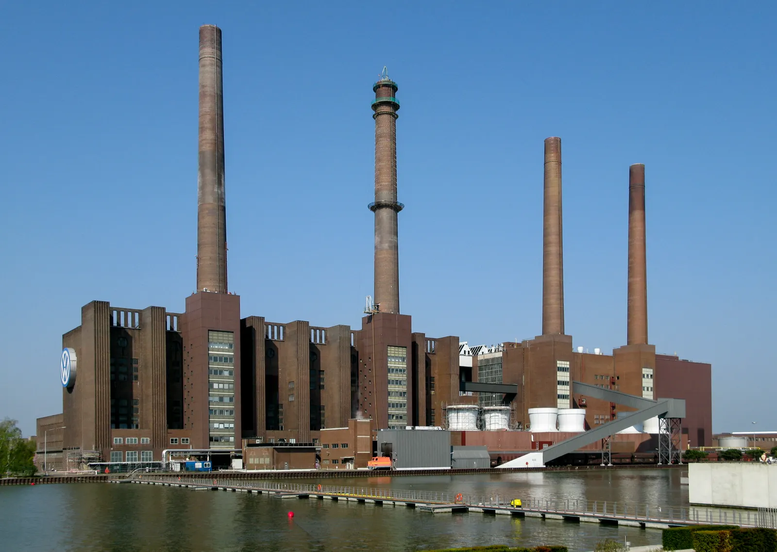 Edificio de ladrillo de la central térmica de la fábrica de Volkswagen en Wolfsburgo, con cuatro chimeneas y el logotipo de la marca, visto desde el canal.